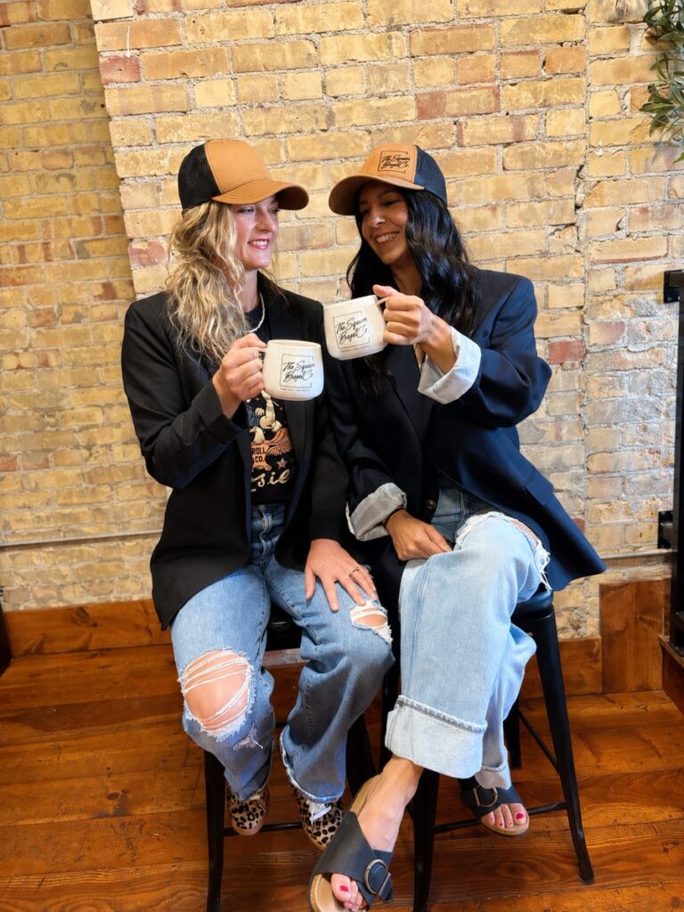 Syd and Alana toasting with Square Bagel Co. mugs in front of the shop’s brick wall